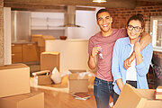 a young couple unpack their belongings as they settle into their new loft apartment . They are hugging and looking to camera smiling holding their keys aloft .