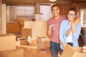 a young couple unpack their belongings as they settle into their new loft apartment . They are hugging and looking to camera smiling holding their keys aloft .